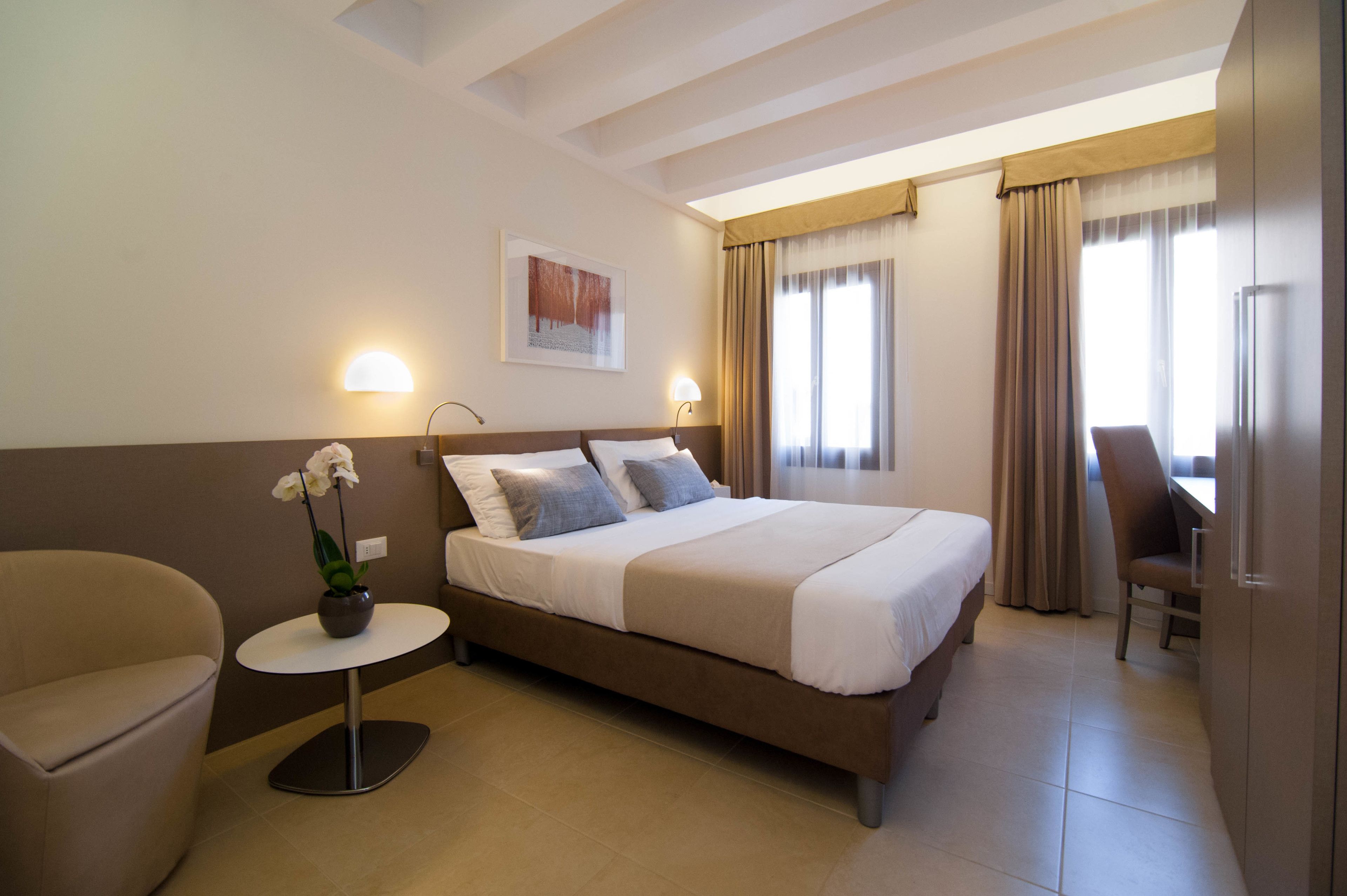 A hotel room with a bed, a chair, a small table with a flower, and a desk with a chair.