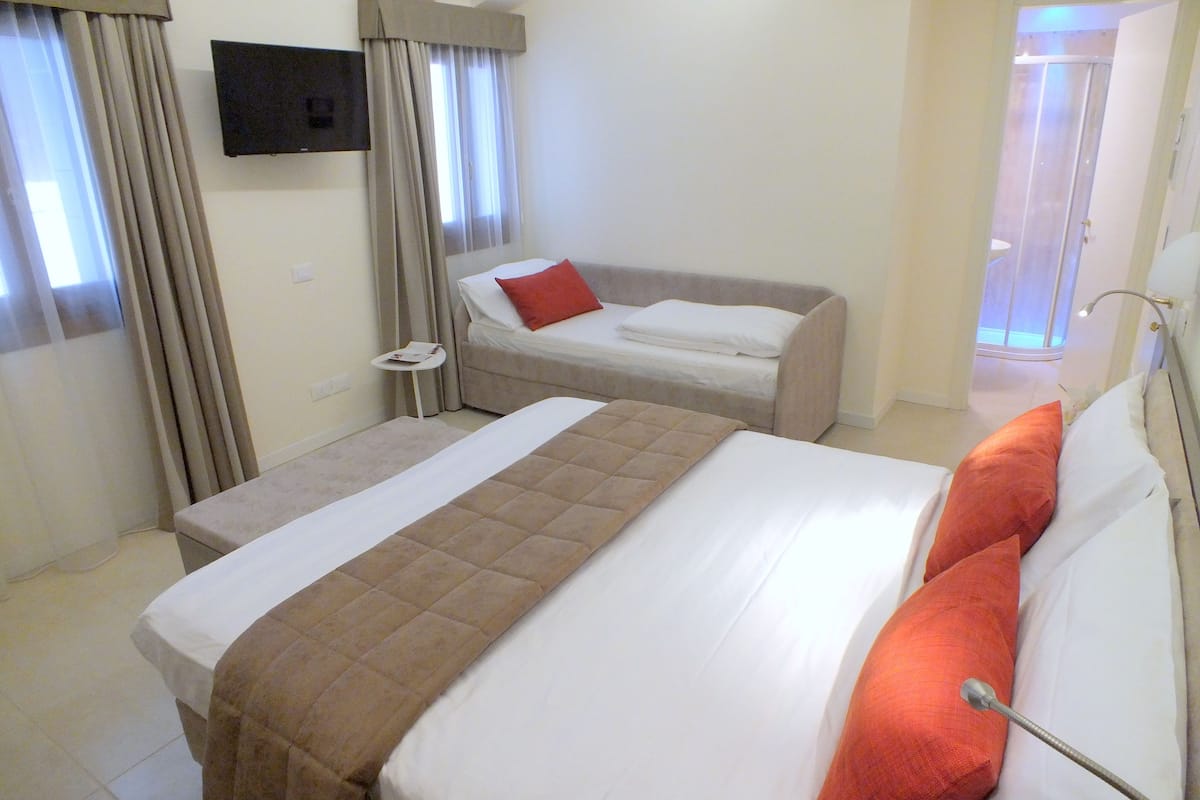 Hotellihuoneessa on suuri sänky, pienempi sänky, televisio ja kylpyhuone, joka näkyy avoimesta ovesta.