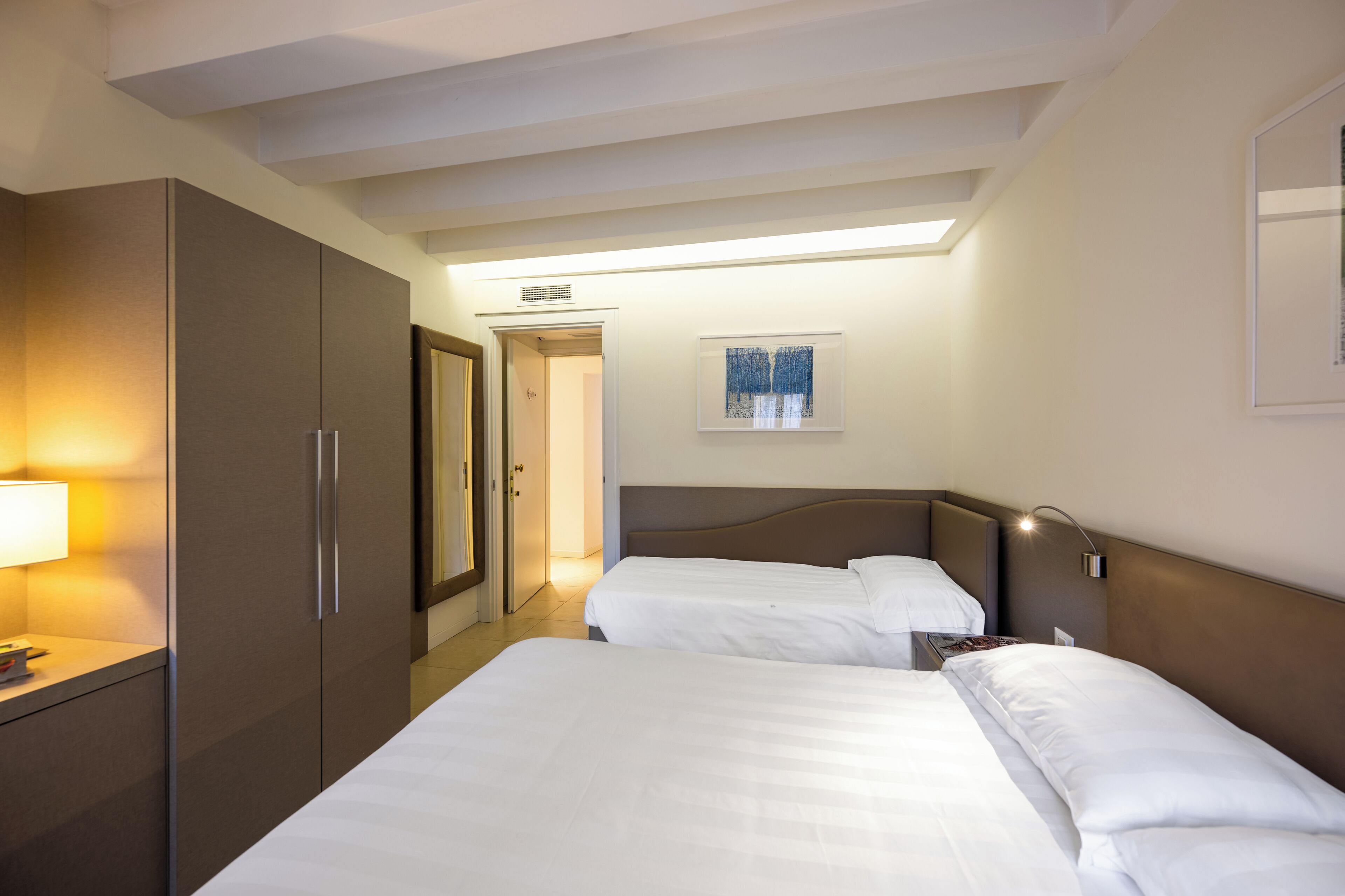 A hotel room with two beds, a wardrobe, a bedside table, and a wall-mounted lamp.
