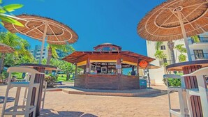 A central wooden kiosk with a sign, surrounded by thatched umbrellas and outdoor seating. - Tsokkos Gardens (Paralimni)