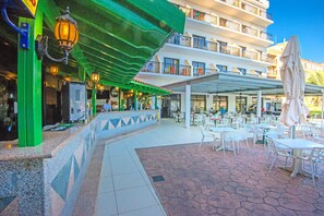 An outdoor cafe area with tables and chairs, a covered seating area, and a building with balconies in the background. - Tsokkos Gardens (Paralimni)