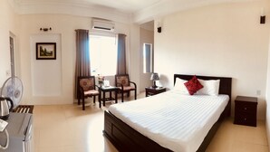 A hotel room with a bed, a desk with a chair, a TV, a fan, a kettle, and a telephone. - Nam Long Plus Hotel (Dong Hoi)