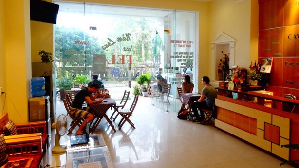 A cafe with a modern interior, featuring a reception desk, seating area with tables and chairs, and large windows providing a view of the outdoors. - Nam Long Plus Hotel (Dong Hoi)