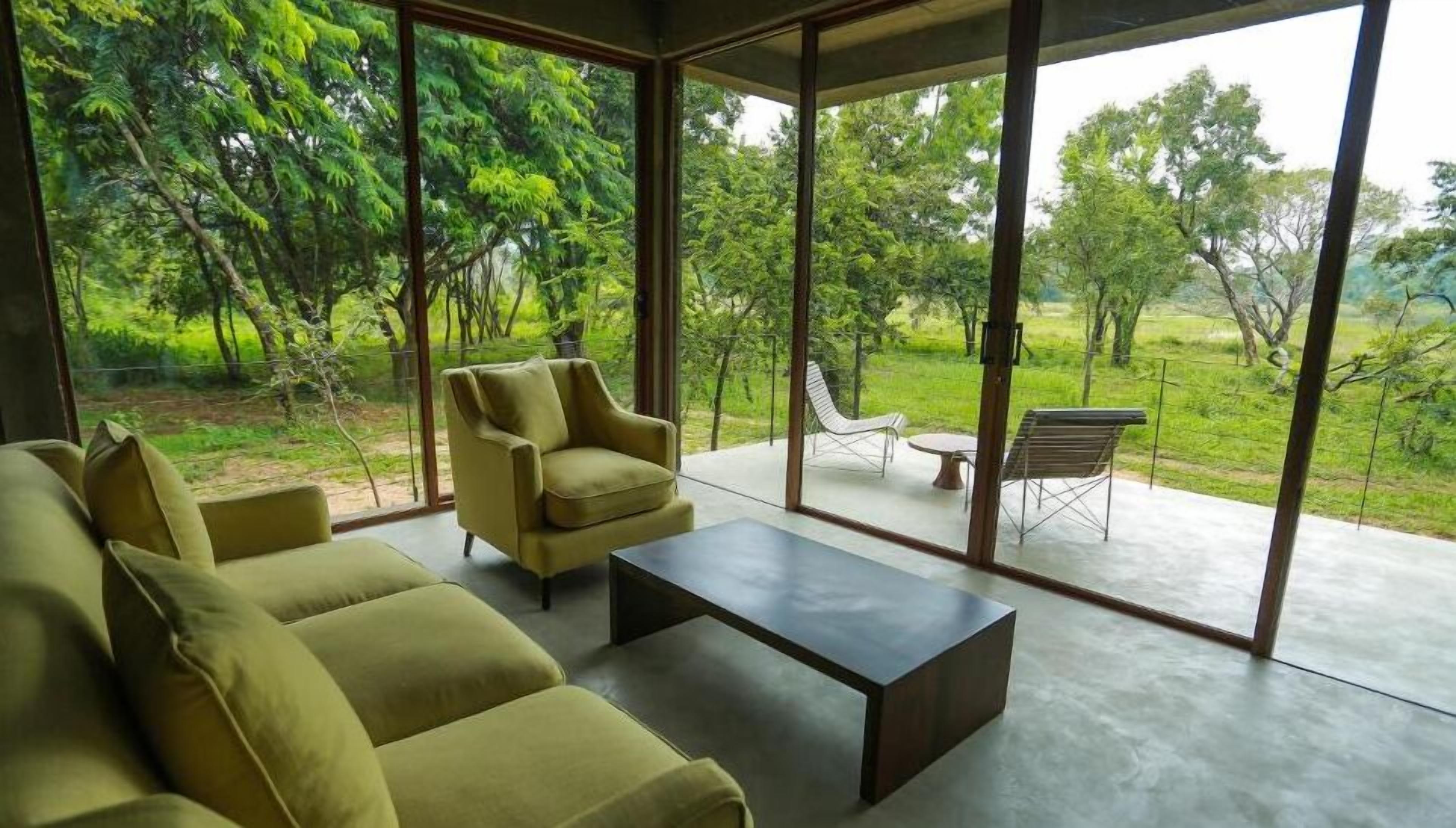 A modern living room with a sofa set and a coffee table, large glass doors leading to an outdoor patio with a view of a lush garden.