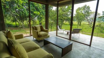 A modern living room with a sofa set and a coffee table, large glass doors leading to an outdoor patio with a view of a lush garden.