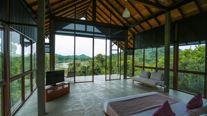 A spacious room with a large bed, a sofa, and a TV on a wooden stand. The room has large windows offering a view of lush greenery and hills. - Wild Grass Nature Resort (Dambulla)