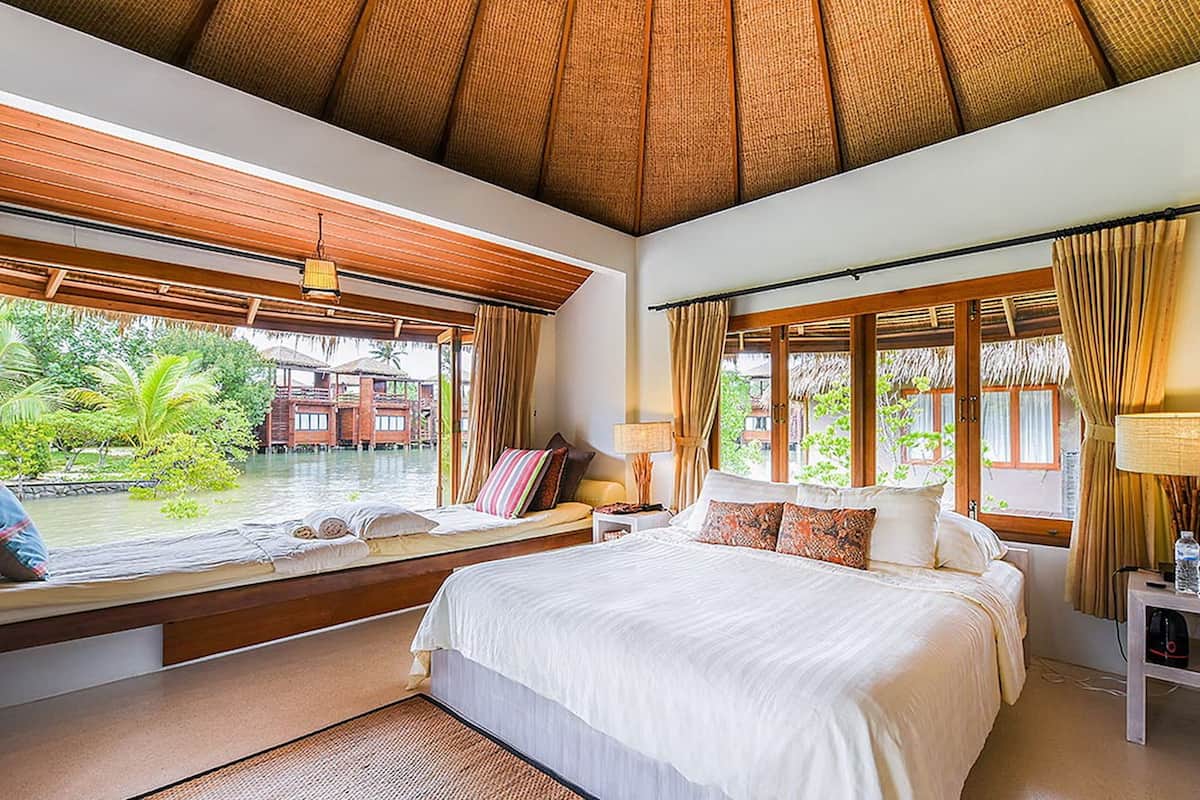 Un dormitorio amplio con una cama grande, un asiento junto a la ventana con vistas a un cuerpo de agua y una vista de una estructura de madera.