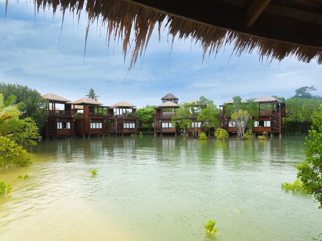 A series of wooden overwater bungalows with thatched roofs, surrounded by calm water and lush greenery.