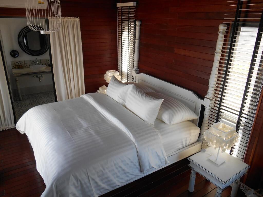 A neatly made bed with white linens in a room with wooden walls and a window with blinds.