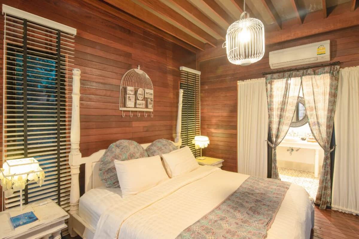 Un dormitorio acogedor con techo de madera, una cama con ropa de cama estampada, una mesita de noche con lámpara y una ventana con cortinas.