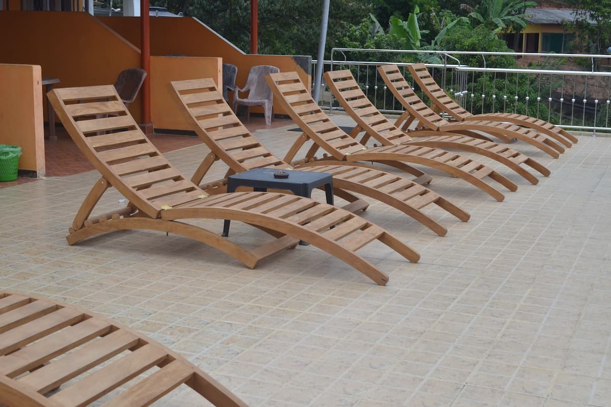 A row of wooden lounge chairs with a small table in between, arranged on a tiled patio area.