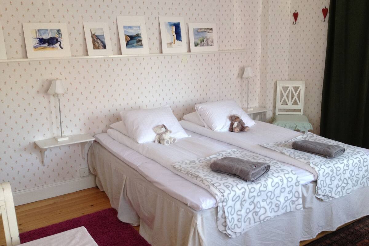 A double bed with white bedding and a patterned duvet, a bedside table with a lamp, a small shelf with framed pictures, and a red curtain.