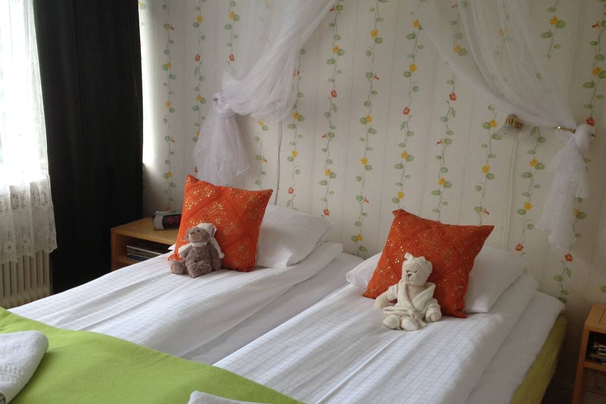 A double bed with white linens, two orange pillows, and a green throw blanket.