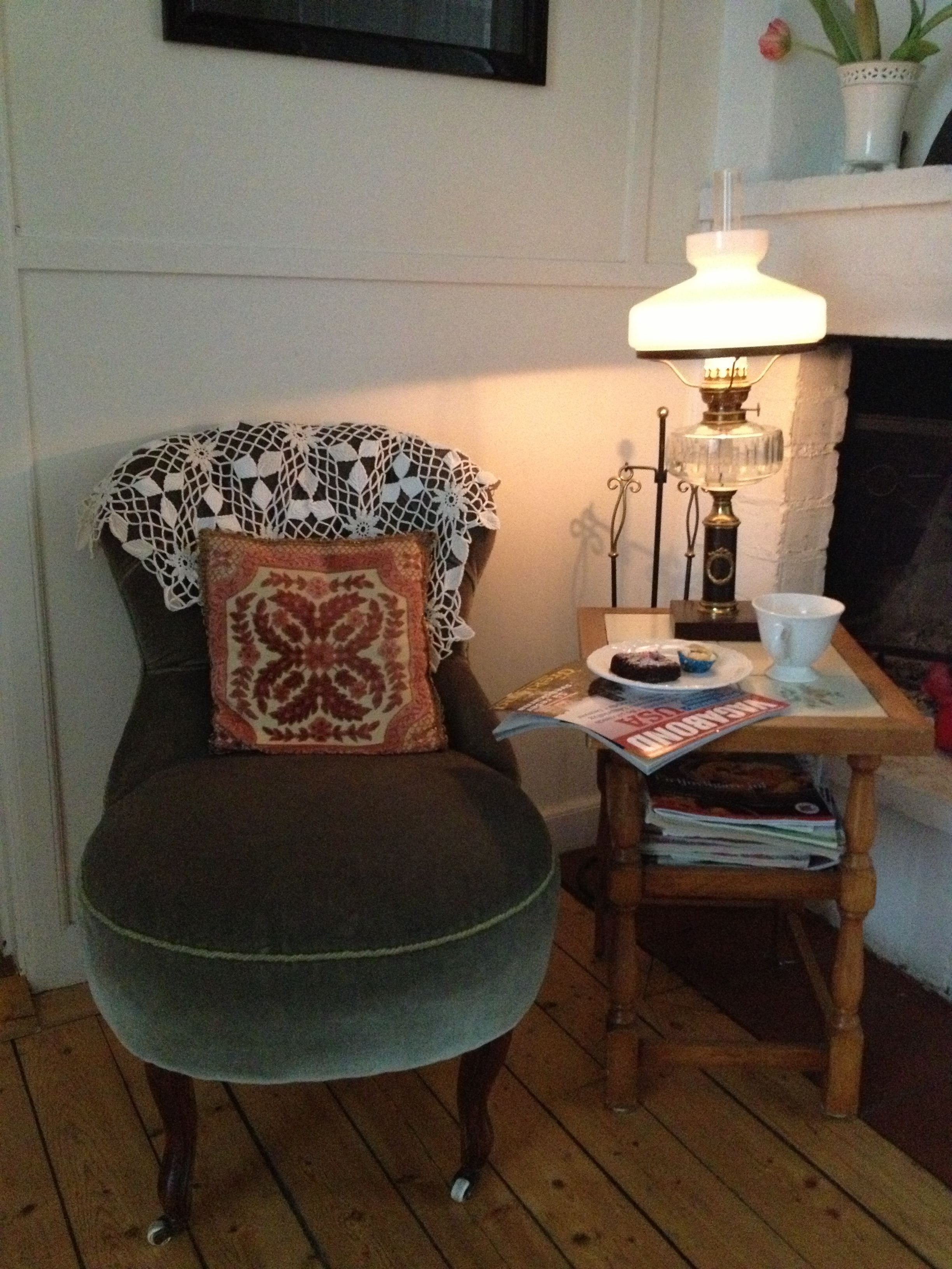 Una sala de estar acogedora con un sillón verde oscuro, un chal de encaje, un cojín estampado, una mesita de madera pequeña con una lámpara, un plato de comida y una revista.