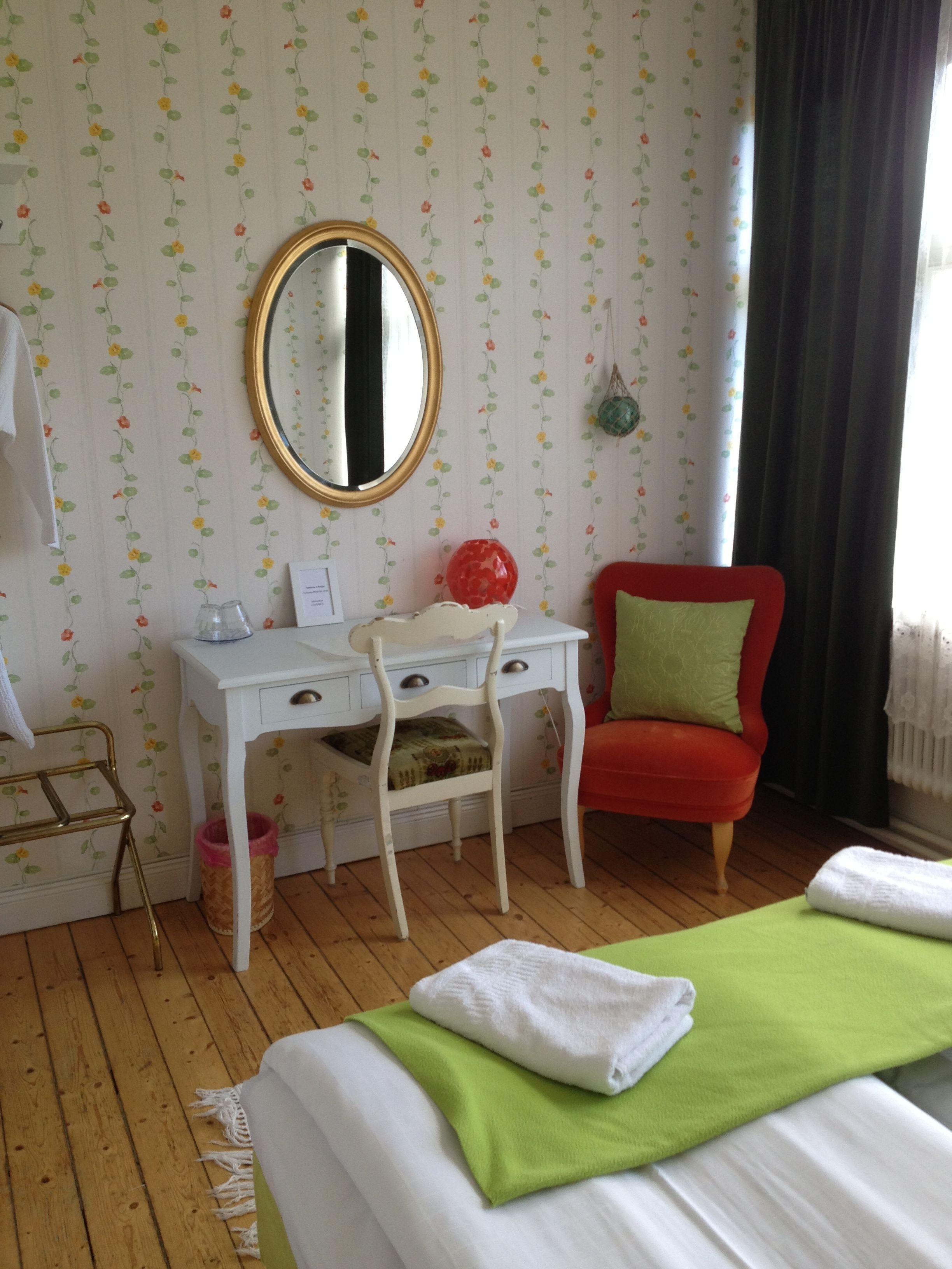 Un dormitorio con cama, un escritorio con silla, una silla roja, un espejo y una ventana con cortinas.