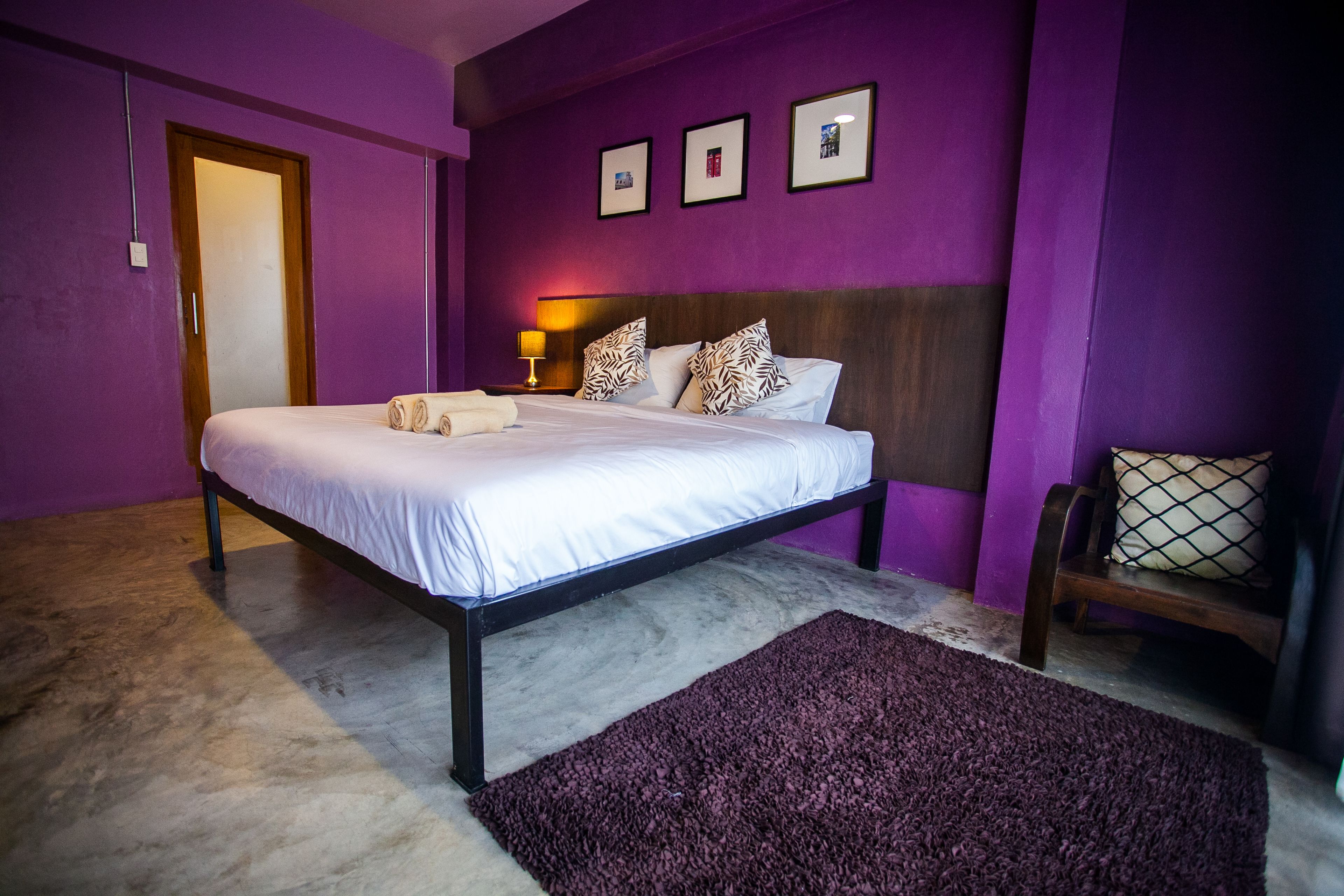 Un dormitorio con una cama, cabecera de madera, pared morada y una mesita de noche con una lámpara.