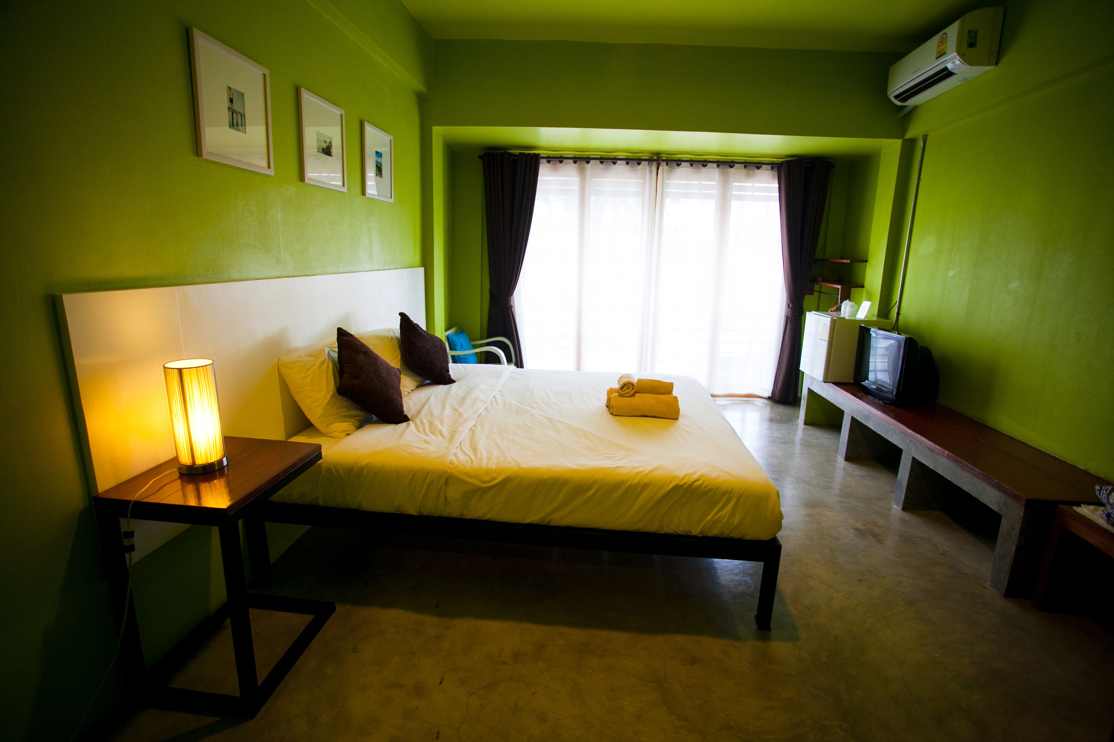Una habitación con cama, mesita de noche, lámpara y televisor sobre una banca.