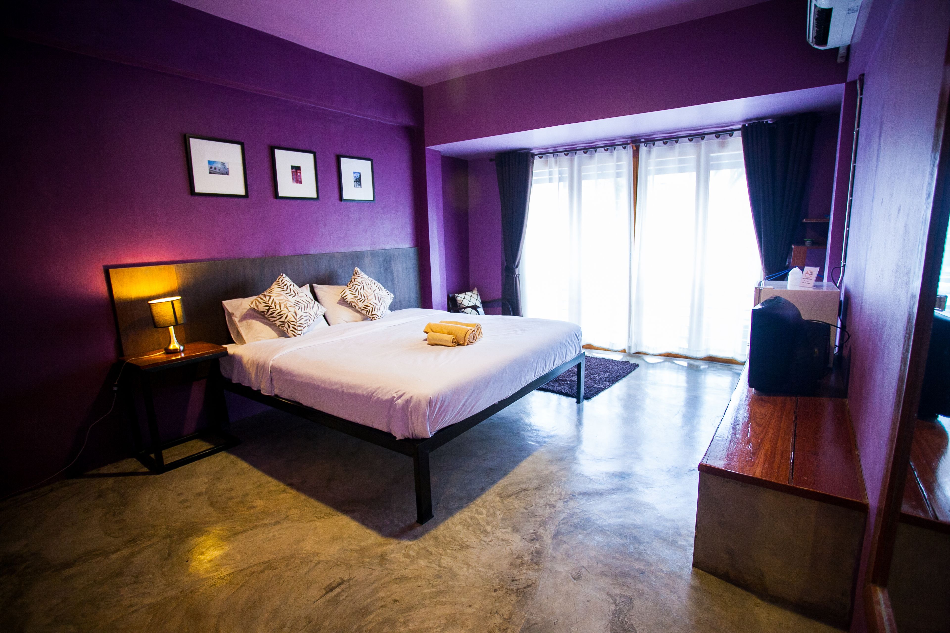 A bedroom with a bed, a nightstand, a lamp, a window with curtains, and a small table with a chair.