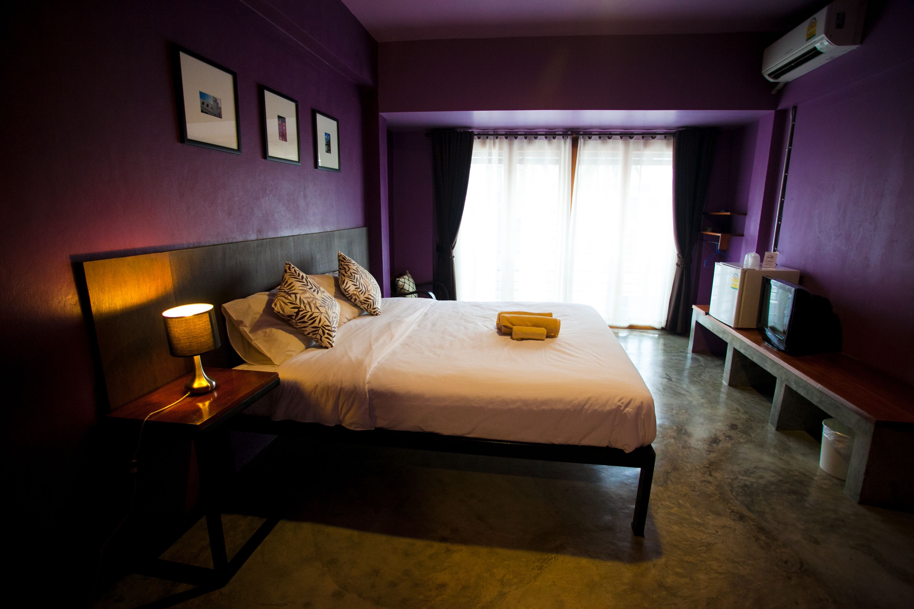 A bedroom with a bed, bedside lamp, air conditioning unit, and a television on a bench.