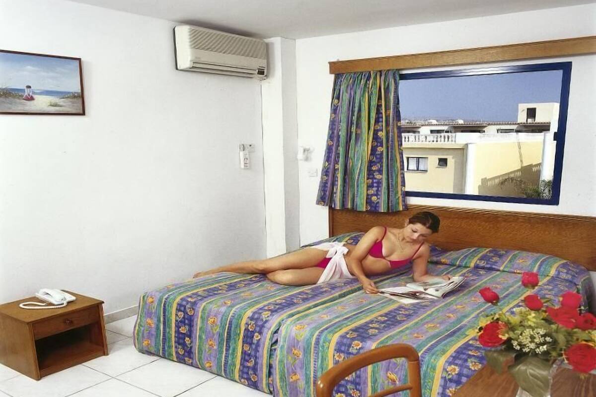 A single bed with a colorful floral bedspread, a wooden headboard, a small bedside table with a telephone, and a window with curtains.