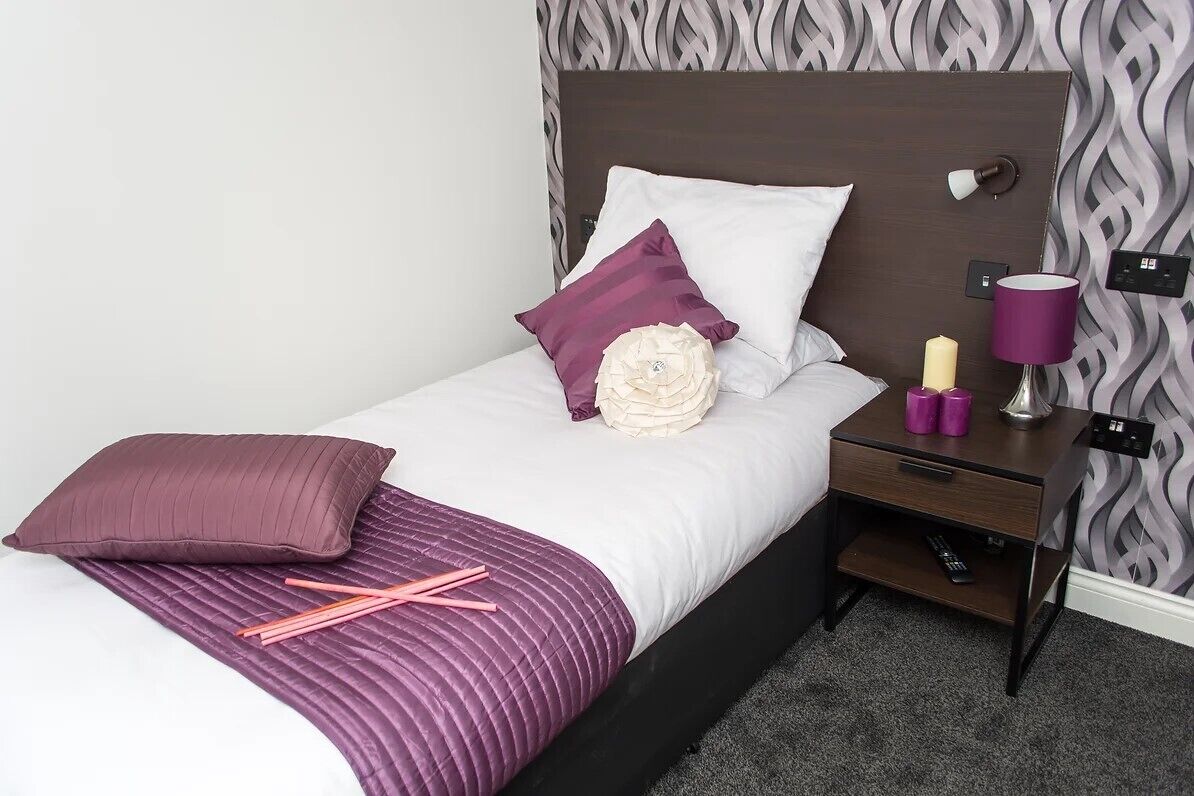 Ein ordentlich bezogenes Bett mit einer weißen und violetten Tagesdecke, zwei Kissen und einer Zierblume. Ein Nachttisch mit Lampe, Fernbedienung und einer kleinen Schublade steht daneben. Der Raum hat einen Wandtapetenmuster und ein dunkles Kopfteil.