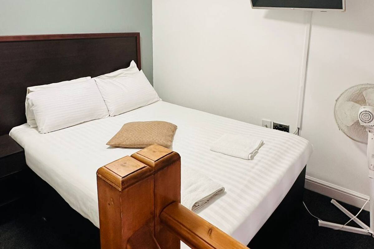 A hotel room with a bed, a TV mounted on the wall, a ceiling light, and a floor fan.