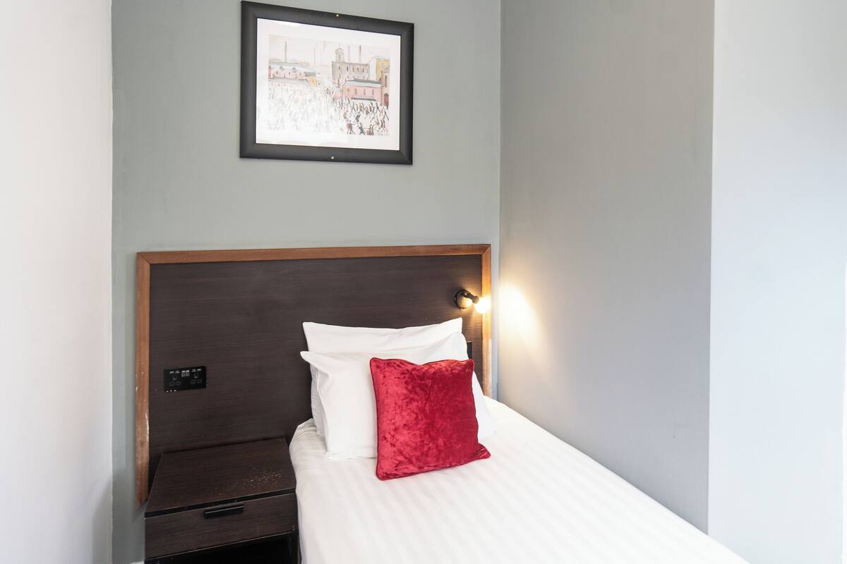 A single bed with a wooden headboard, white bedding, and a red pillow. There is a nightstand with a lamp on it, and a framed picture above the bed.