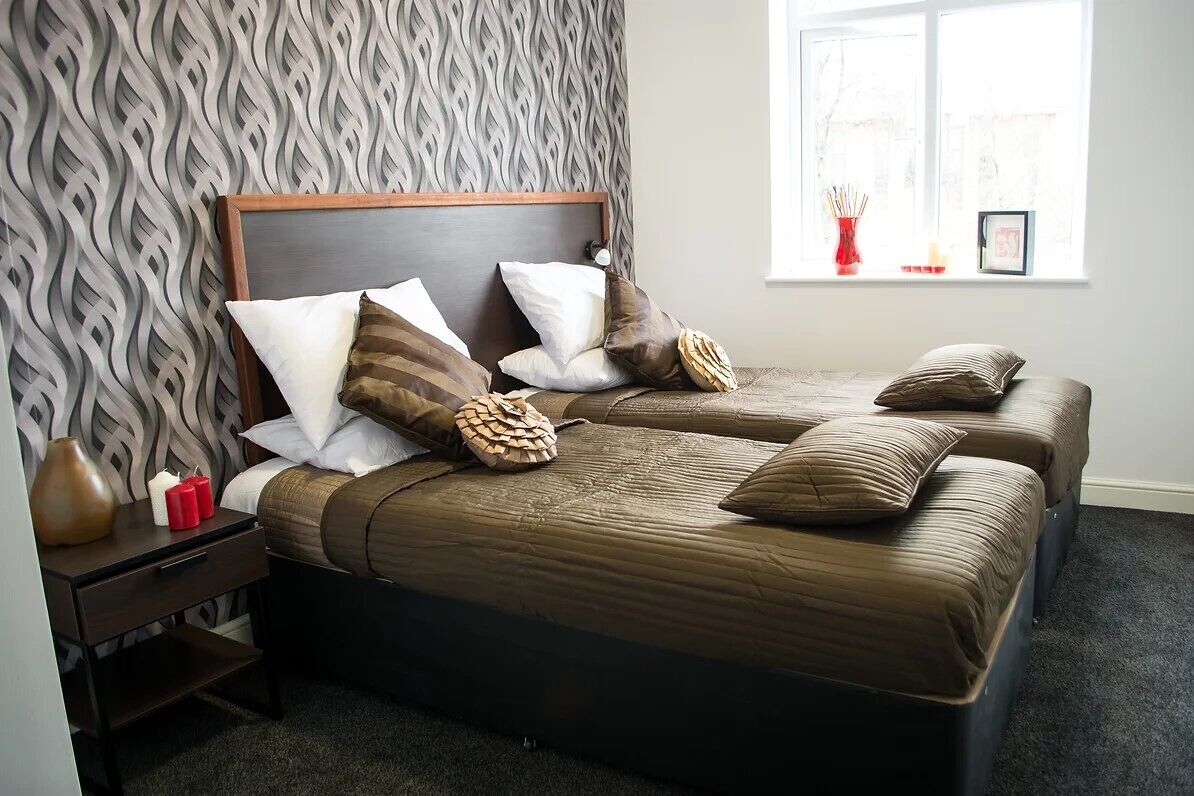 A hotel room with two beds, a patterned wallpaper, a nightstand with a vase, and a window with a view.