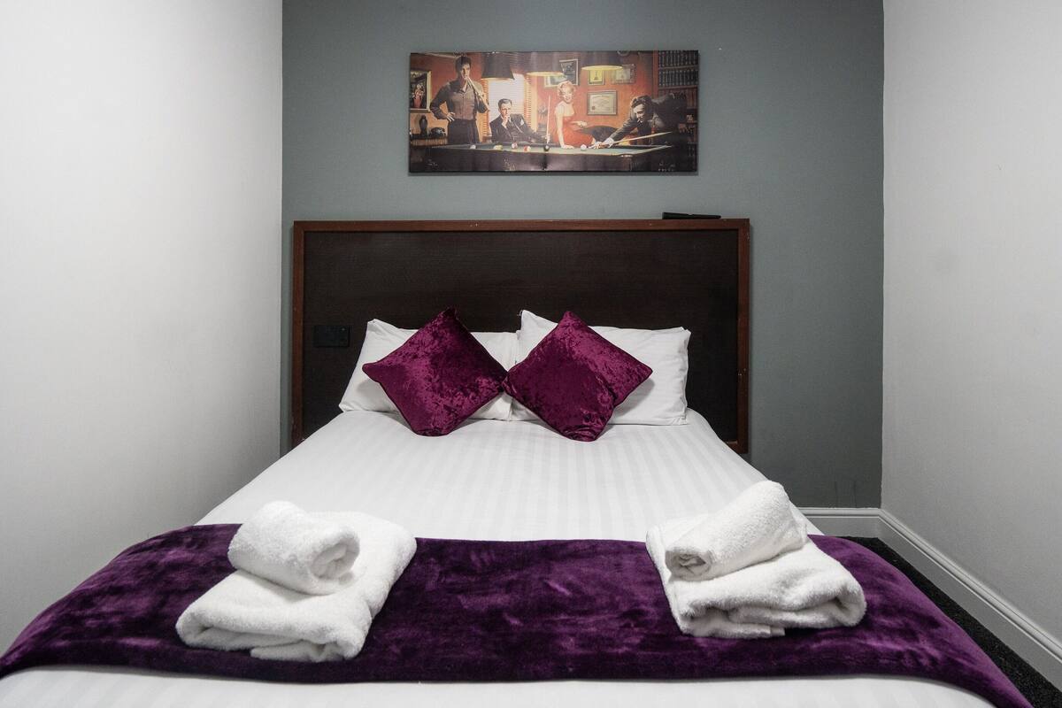 A neatly made bed with white linens, two purple pillows, and two folded white towels.