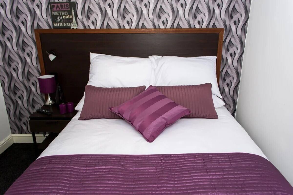 A neatly made bed with white and purple bedding, a wooden headboard, and a patterned wallpaper with a sign above it.