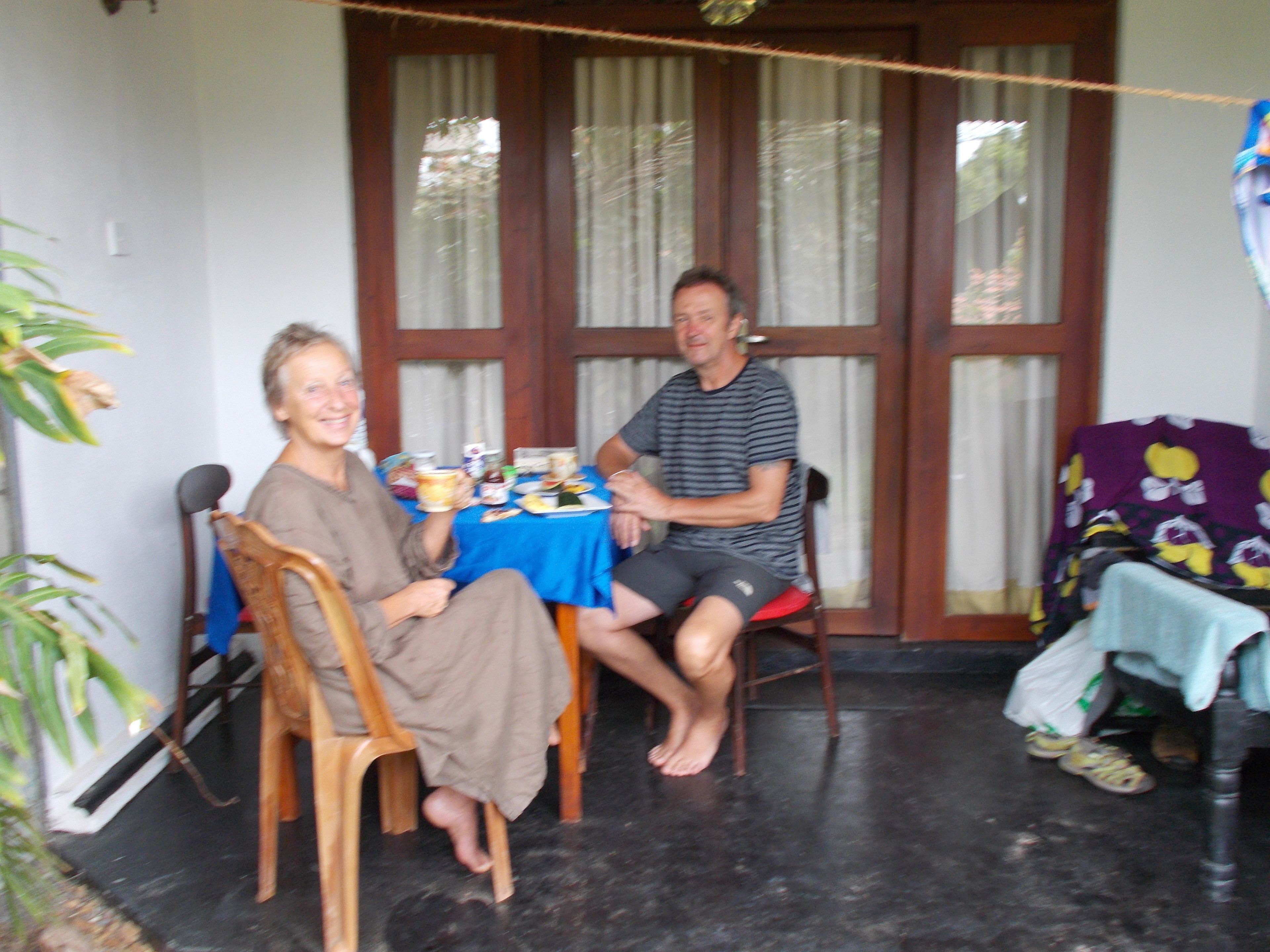 Two people seated at a table with a blue tablecloth, wooden chairs, and a window with curtains.