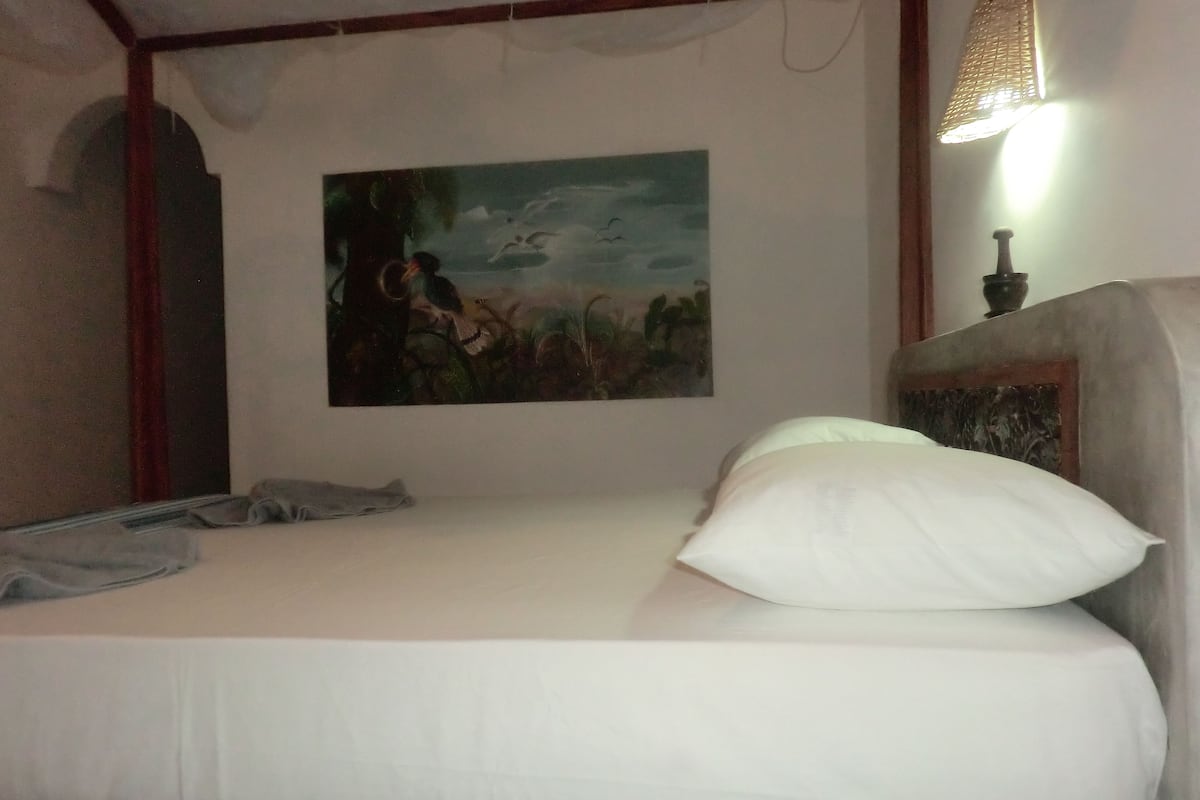 A bedroom with a bed, a painting on the wall, and a lamp.