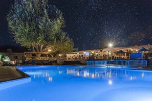 A swimming pool with a starry night sky above, surrounded by a building with outdoor seating and a bar. - Gelli Apartments (Kos)