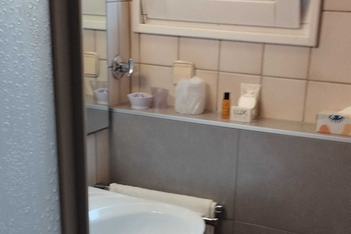 Une salle de bain avec un lavabo blanc, des toilettes, une petite poubelle et une fenêtre au-dessus du lavabo.