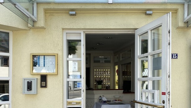 L’entrée d’un hôtel, avec une enseigne indiquant « HÔTEL » au-dessus de la porte. On y trouve un petit comptoir d’accueil avec un guichet et une chaise. L’entrée est précédée d’un petit escalier.