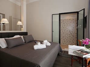 A hotel room with a bed, a shower, a table with a book and a vase of flowers, and a mirror with lamps. - Residenza Cavour (Rome)