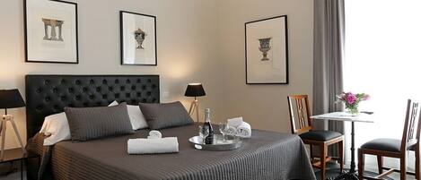 A hotel room with a large bed, a bedside table with a lamp, a small table with a vase of flowers, and framed artwork on the walls.