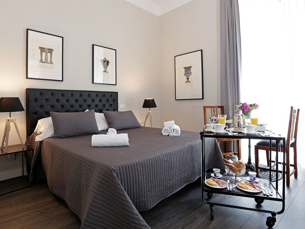 A hotel room with a large bed, a bedside table with a lamp, a breakfast trolley with food and drinks, and framed pictures on the wall.