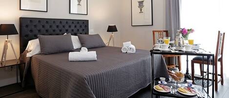 A hotel room with a large bed, a bedside table with a lamp, a breakfast trolley with food and drinks, and framed pictures on the wall.