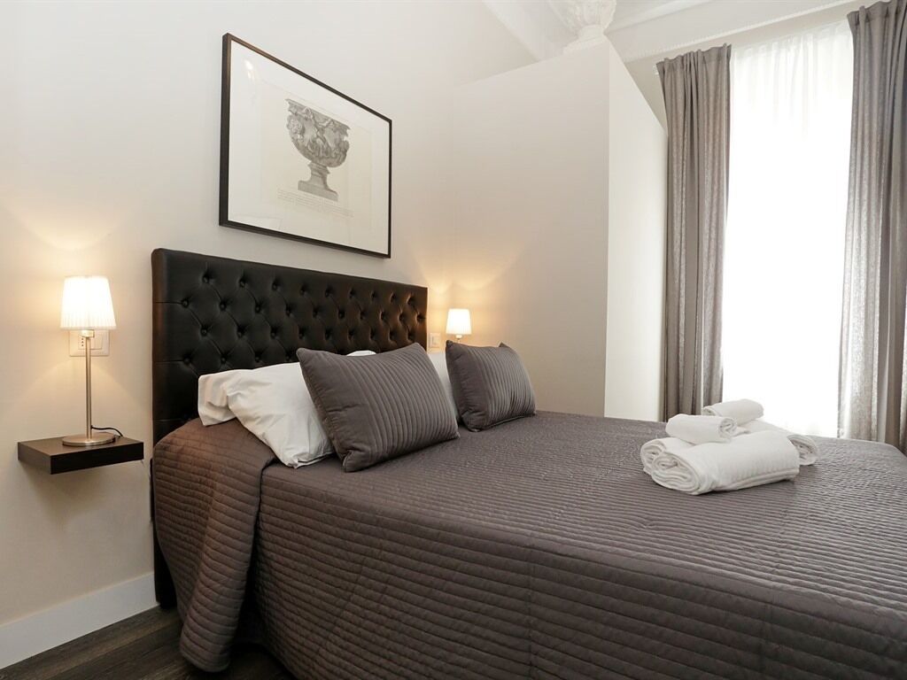 A neatly made bed with a dark headboard, two bedside lamps, a framed picture on the wall, and a window with curtains.