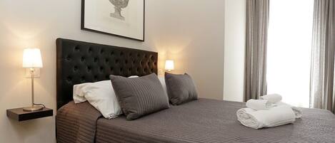 A neatly made bed with a dark headboard, two bedside lamps, a framed picture on the wall, and a window with curtains.