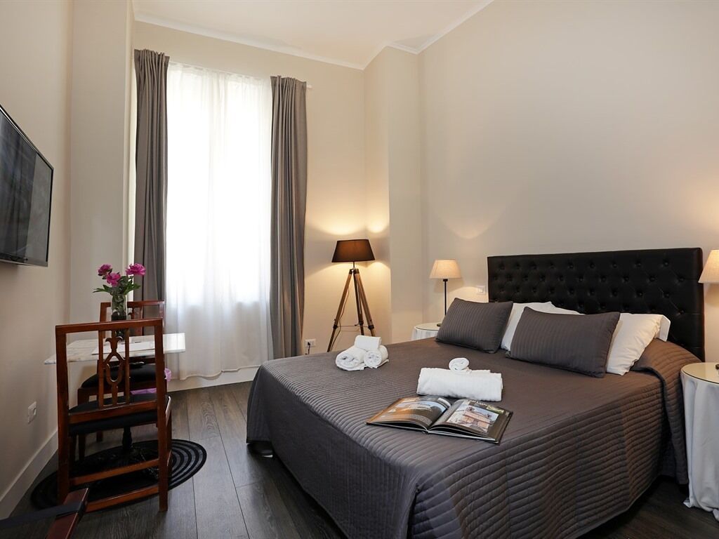 A hotel room with a bed, bedside tables, a lamp, a chair, a desk, a TV, and curtains.