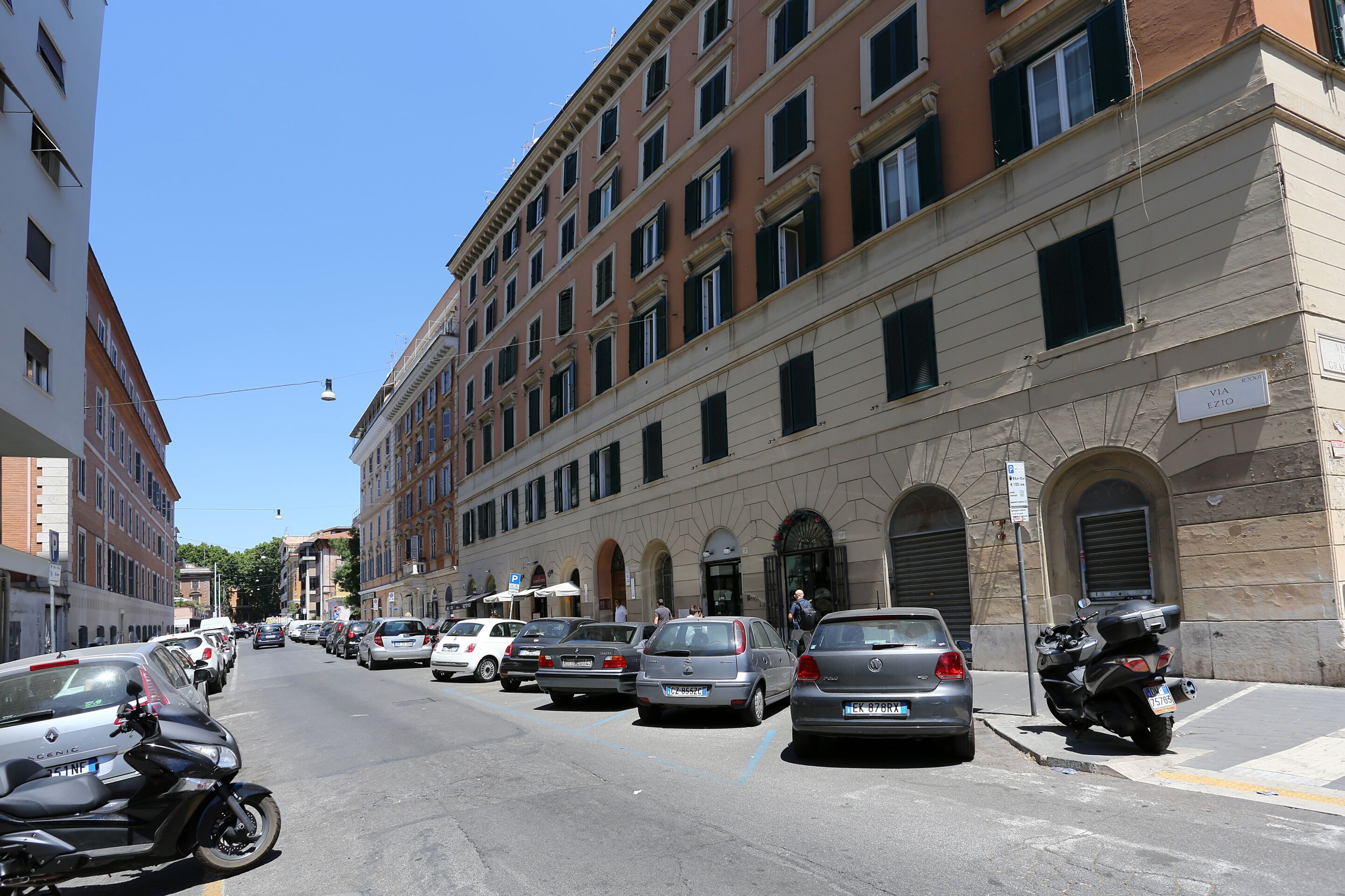 Une rue bordée de voitures garées et d’une moto, des immeubles dotés de nombreuses fenêtres et un ciel dégagé.