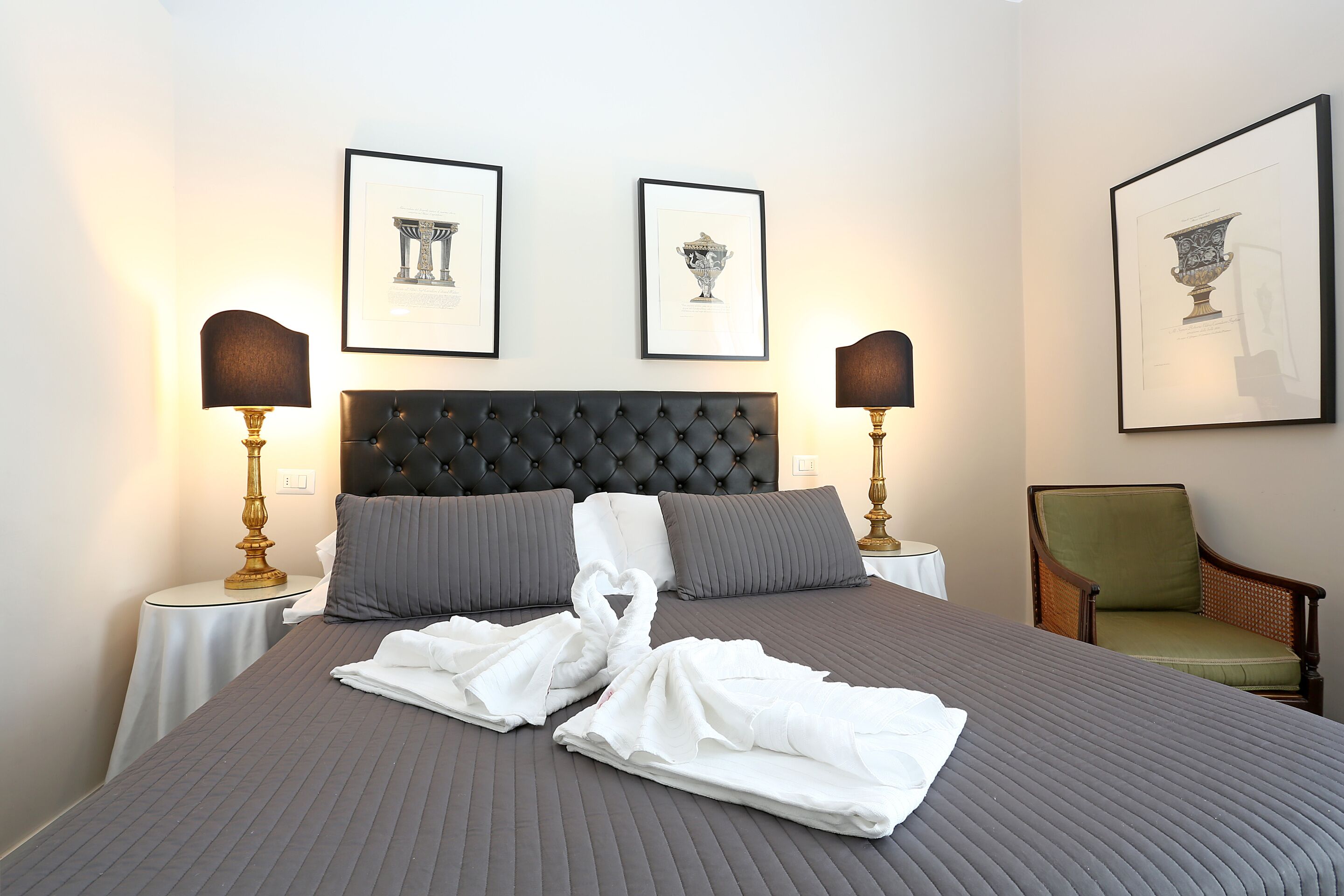 Una cama bien tendida con una colcha gris, dos almohadas y dos toallas dispuestas en forma de corazón. En la pared, encima de la cama, hay cuadros enmarcados, y se ve una silla en la esquina.