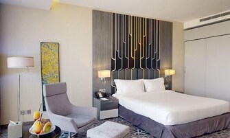 A modern hotel room with a large bed, a side table, a chair, a floor lamp, and a wall with a geometric design.