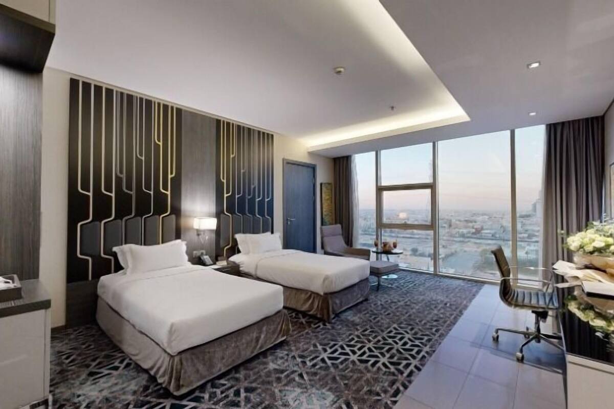 A modern hotel room with two beds, a desk, and a large window offering a city view.