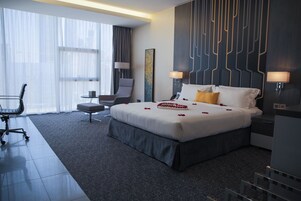 Habitación de hotel moderna con una cama grande, un escritorio, una silla y vistas de la ciudad a través de cortinas translúcidas.