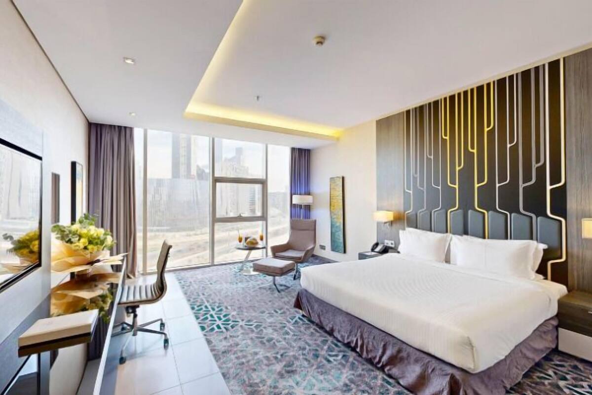 A modern hotel room with a large bed, a desk with a chair, a small table with a vase of flowers, and a view of the city through large windows.