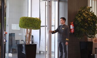 A hotel reception area with a receptionist standing by a glass door, a potted plant, and a fire extinguisher on the wall.