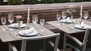 A restaurant setting with wooden tables, white plates, and wine glasses, set against a backdrop of neatly trimmed bushes.